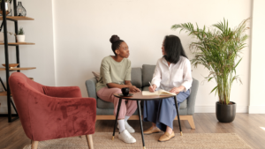 person with dark complexion and braids looking at a person with light complexion and dark hair. both are seated on a couch in a therapists office